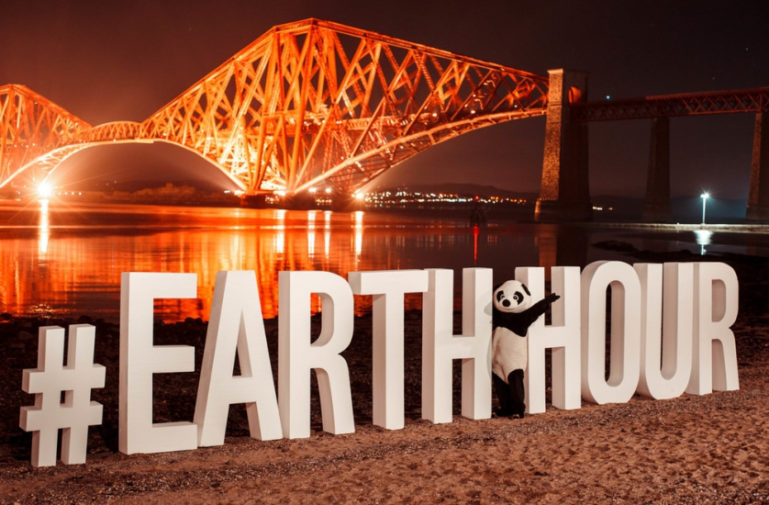 Photo du logo en 3D de Earth Hour avec une personnage portant un costume de panda devant un pont allumé de nuit