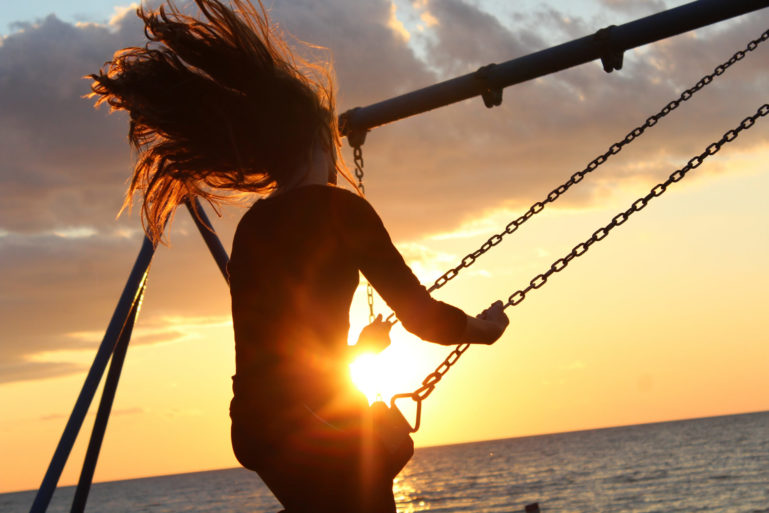Photo de dos d'une jeune fille au bord de la mer faisant de la balançoire les cheveux au vent, devant un soleil couchant 