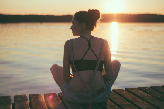 Photo d'une jeune fille de dos en assise au bord d'un ponton avec un soleil couchant à l'horizon